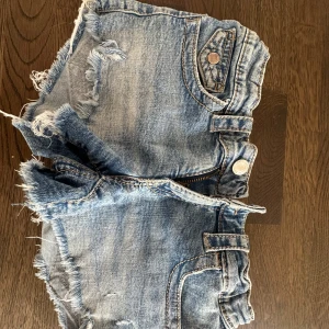 Blå jeansshorts från Gina Tricot - Snygga blå jeansshorts från Gina Tricot med fransiga kanter och slitna detaljer. Shortsen har fickor både fram och bak med lock och knapp, samt klassisk jeansknapp och dragkedja. Perfekta för varma dagar!I storlek 152
