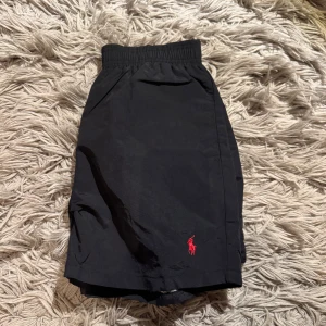 Polo Ralph Lauren badbyxor - Svarta badshorts från Polo Ralph Lauren med röd logga broderad på benet. Elastisk midja med röd insida och innerbyxa i mesh. Perfekt nu till sommar väder! Aldrig använd. 10/10 skick. Storlek är M men passar S. Priset är ej hugget i sten. Bara att skriva vid minsta funderingar!