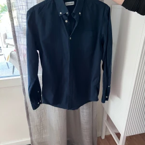 Mörkblå skjorta från Jack & Jones - Snygg mörkblå skjorta från Jack & Jones med klassisk krage och knappar framtill. Skjortan har lång ärm och en bröstficka, perfekt för en stilren look. Passar till många olika tillfällen.