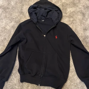 Svart hoodie från Ralph Lauren - Svart hoodie med dragkedja från Ralph Lauren. Klassisk design med huva, fickor framtill och röd broderad logga på bröstet. Perfekt för en avslappnad stil.