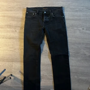 Svarta jeans från Nudie Jeans - Säljer ett par svarta jeans från Nudie Jeans med klassisk femficksdesign och raka ben. Jeansen har silverfärgade nitar och knapp, samt diskret logga på bakfickan. Perfekta för dig som gillar stilrena och tidlösa jeans.