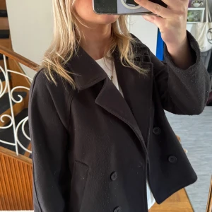 Svart kappa från H&M - Säljer en svart, kort kappa från H&M med dubbelknäppning framtill och klassisk krage. Har varit använd en del men ändå i helt okej skick!