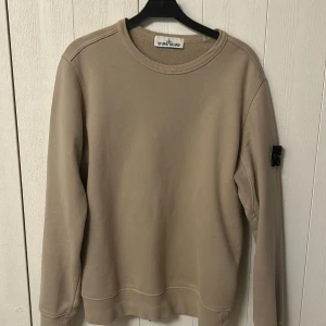 Beige sweatshirt från Stone Island - Säljer en beige sweatshirt från Stone Island med klassisk logotyp-patch på ärmen. Tröjan har rund halsringning och långa ärmar. Collagetröja beige. Storlek M