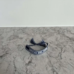 Blått armband från Christian Dior - Snyggt blått armband från Christian Dior med vit text och tofsar i ändarna. Armbandet är justerbart och har texten 'Christian Dior J'Adior' broderat. Perfekt accessoar för att lyfta din stil.