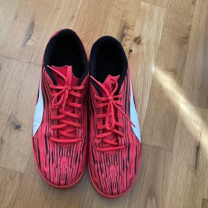 Röda fotbollsskor från Puma - Säljer ett par röda fotbollsskor från Puma med svart och vitt mönster. Skorna har snörning och en vit Puma-logga på sidan. Yttersulan är mönstrad för bra grepp på planen. Perfekta för dig som vill sticka ut på fotbollsplanen!