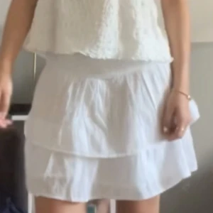 Vit volangkjol från Gina Tricot - Säljer en söt vit kjol från Gina Tricot med bred resår i midjan och dubbla volanger. Kjolen är kort och har ett broderat mönster i tyget som ger en romantisk känsla. Perfekt till varma dagar och enkel att matcha med olika toppar.