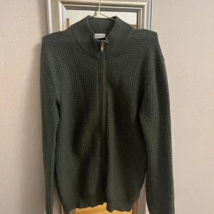 Mörkgrön stickad kofta från John Henric - Säljer en mörkgrön stickad kofta från John Henric i storlek M. Koftan har hel dragkedja framtill, hög krage och ett diskret rutmönster i stickningen. passar till många tillfällen.
