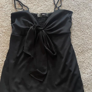 Svart linnetopp med rosett från ONLY - Säljer en svart linnetopp från ONLY med smala axelband och en snygg rosettdetalj framtill. Toppen har en smockad rygg för extra komfort och sitter snyggt på kroppen. Perfekt till sommarens alla tillfällen!