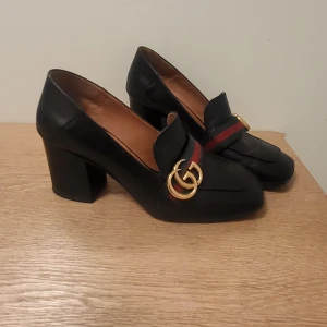 Svarta klackloafers från Gucci - Säljer ett par svarta loafers med klack från Gucci. Skorna har rund tå, bred klack och är dekorerade med det klassiska GG-spännet i guld samt röd-gröna band över vristen. Perfekta för dig som vill ha en lyxig och tidlös look.