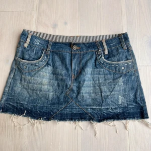 Jeanskjol med fransig kant - Säljer en blå jeanskjol med fransig nederkant och klassiska fickor fram och bak. Kjolen har bälteshällor, knapp och dragkedja framtill samt dekorativa nitar på fickorna. Perfekt för en avslappnad stil.