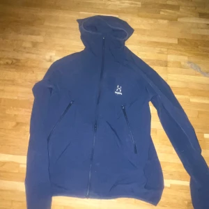 blå haglöfs jacka - Mörkblå hoodiejacka från Haglöfs med hel dragkedja och två snedställda fickor framtill. Jackan har huva och är tillverkad i ett mjukt syntetmaterial, perfekt för lager-på-lager eller som lättare ytterplagg.