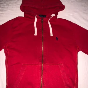 Röd hoodie från Polo Ralph Lauren - Röd hoodie från Polo Ralph Lauren med dragkedja och vit snörning i huvan. Klassisk broderad logga på bröstet och två fickor framtill. Perfekt för en avslappnad stil. Finns litet hål inne vid luvan men inget som syns vid användning.