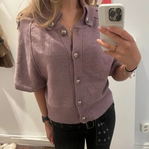 Ljuslila stickad kofta med kort ärm och knappar - Säljer en ljuslila stickad kofta med korta ärmar och stora silvriga knappar framtill. Koftan har bred krage och ribbade muddar i nederkant. Perfekt att styla till både jeans och kjol. Storlek L men sitter bra på mig som har S i vanliga fall.