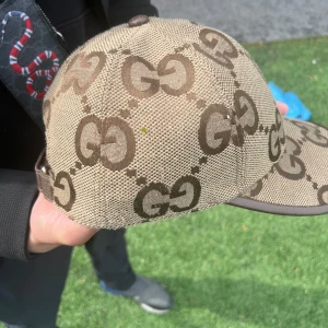 Beige keps från Gucci med GG-mönster - Säljer en beige keps från Gucci med det klassiska GG-mönstret i brunt. Kepsen har böjd skärm och är tillverkad i ett slitstarkt material. Perfekt för dig som vill ha en stilren och lyxig accessoar.