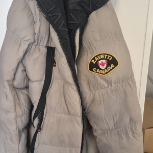 Beige pufferjacka från Zavetti Canada - Säljer en beige pufferjacka från Zavetti Canada med svart innerfoder och coolt märke på ärmen. Jackan har dragkedja framtill, praktiska fickor och en rejäl krage. Perfekt för kalla dagar när du vill hålla stilen och värmen. Dock lite skitig så måste tvättas