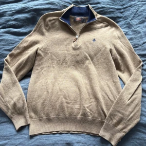 Beige stickad half zip-tröja Morris - Snygg beige stickad tröja från Morris med half zip och mörkblå insida på kragen. Broderad logga på bröstet och ribbade muddar. 