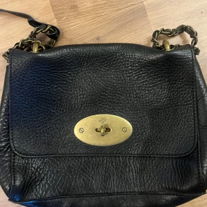 Mulberry ”Lilly” axelväska i skinn - Säljer en klassisk svart axelväska från Mulberry i grymt snyggt skinn med kedjerem och ikoniskt vridlås i mässing. 21 cm bred (max 25 cm) x 17 cm hög x 9 cm djup Axelremmen: 28 cm när den är dubbel och 53 cm enkel. Låset fattas och den är i fint skick men i använt skick. Kvitto och dustbag saknas tyvärr, den köptes för många år sedan. Och därav billigare än andra som är ute för försäljning. 