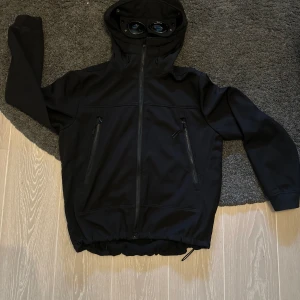 Svart Goggle Jacket från C.P. Company - Svart Goggle Jacket från C.P. Company med ikoniska glasögon i huvan. Jackan har dragkedja framtill, två snedställda fickor med dragkedja och justerbar nederkant. Tillverkad i mjukt syntetmaterial med fleecefoder för extra komfort. Perfekt för dig som gillar streetwear med edge. bra till hösten. storlek M passar folk runt 180cm nypris 4799kr