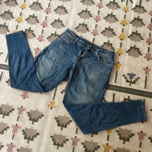 Blå Nudie Jeans - Säljer ett par klassiska blå jeans från Nudie i storlek W31 L32 i modellen skinny lin. Finns en skavank i grenen (se bild 3), därav priset. De sitter ganska brett och är tillverkade i mjukt jeanstyg. 