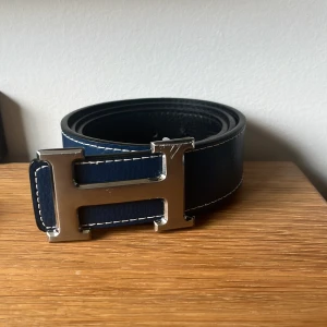 Blått skärp med H-spänne - Bälte med ikoniskt H-spänne i silverfärgad metall. Bältet är gjort i skinn med vit kontrastsöm längs kanterna, vilket ger en stilren och exklusiv känsla. Perfekt accessoar för att lyfta din outfit.