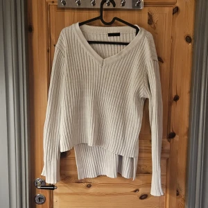 Ribbad vit stickad tröja med v-ringning - Säljer en ribbad vit stickad tröja med v-ringad hals och lång ärm. Tröjan har en lös passform och cool slits i sidorna som ger en snygg detalj. Perfekt att slänga på sig när det är kyligt ute. Den är längre på ryggen än på framsidan. 
