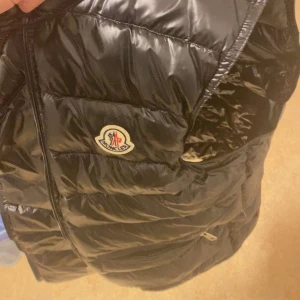 Svart dunväst från Moncler - Snygg svart dunväst från Moncler med glansig finish och klassisk logga på bröstet. Västen har quiltad design och dragkedja framtill. Perfekt att slänga över en hoodie för en cool look.