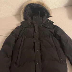 svart Canada goose wyndham black label - Säljer en svart dunjacka från Canada Goose med huva och snygg fuskpäls runt kanten. Jackan har långa ärmar, flera fickor och tydlig logga på ärmen. Perfekt för kalla dagar när du vill hålla dig varm och ändå se stilren ut. Päls ingår självklart och det är storlek M