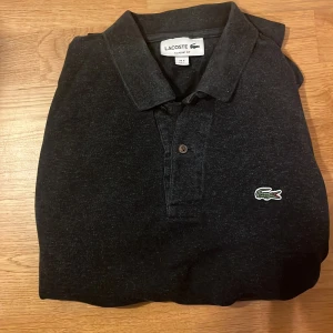 Svart långärmad pikétröja Lacoste - Klassisk svart långärmad pikétröja från Lacoste i storlek M. Tröjan har krage, knäppning framtill och den ikoniska gröna krokodilloggan broderad på bröstet. Perfekt för en clean och stilren look.