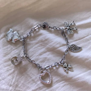 Charmarmband med silverfärgade hängen - Säljer ett charmarmband i silverfärgad metall med flera söta hängen: hjärta, rosett, ring med krona, två fiskar och ett blad. Perfekt för dig som gillar detaljer och vill ha ett unikt smycke med personlig touch.