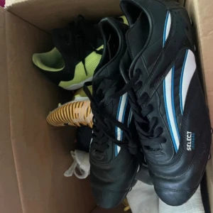 Select svarta fotbollsskor med blå detaljer - Säljer ett par svarta fotbollsskor från Select med vita och blå ränder på sidorna. Skorna har snörning och en robust sula med dobbar för bra grepp på planen. Perfekta för dig som vill steppa upp ditt spel med stil.