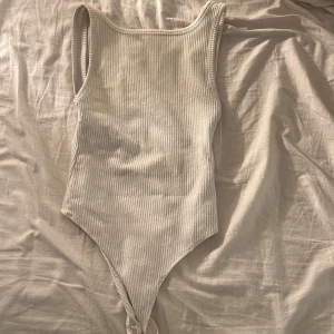 Vit ribbad bodysuit från Bershka - Supersnygg vit ribbad bodysuit från Bershka med ärmlös design och öppen rygg. Gjord i stretchig bomullsmix som sitter tajt och markerar siluetten. Perfekt att styla med jeans eller kjol för en clean look.