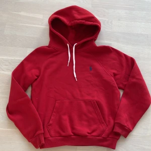 Röd hoodie från Polo Ralph Lauren - Säljer en röd hoodie från Polo Ralph Lauren med klassisk blå logga på bröstet. Tröjan har vit snörning i huvan, stor känguruficka och ribbade muddar. Perfekt för chill dagar och har en skön, mjuk känsla tack vare bomullsmaterialet.