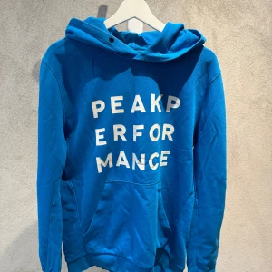 Blå hoodie från Peak Performance M - Säljer en blå hoodie från Peak Performance med stor vit logotyp på framsidan. Tröjan har huva med snörning, magficka och långa ärmar. Materialet är mjuk bomull och insidan är fleecefodrad för extra komfort. Perfekt för chill dagar eller när du vill ha en sportig look.