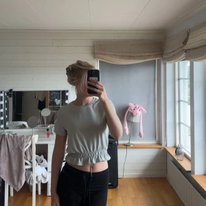 Ljusgrå croppad topp med resår - Säljer en ljusgrå croppad topp med korta ärmar och resår i midjan som ger en snygg rynkad effekt. Enkel och stilren design, perfekt till jeans eller kjol. Superfin att matcha med smycken eller lager på lager.