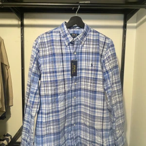 Blårutig skjorta från Polo Ralph Lauren - Snygg blårutig skjorta från Polo Ralph Lauren. Skjortan har klassisk krage, knappar framtill och lång ärm. Tillverkad i mjuk bomull och har den ikoniska broderade loggan på bröstet. Skjortan är helt ny, prislappen är kvar.