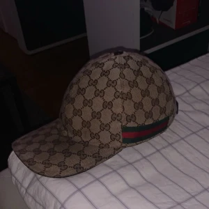 Gucci keps - Säljer en snygg beige keps från Gucci med det klassiska GG-monogrammet över hela kepsen. På sidan finns den ikoniska gröna och röda randen. Kepsen har böjd skärm och justerbar rem baktill. Perfekt för dig som vill ha en lyxig streetstyle-accessoar.