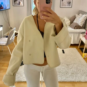 Beige omlottväst från GinaTricot - Säljer en beige omlottväst från Gina med v-ringad hals och vida ärmar. Västen har en croppad passform och stängs med knappar framtill. Perfekt för lager-på-lager och enkel att styla till olika looks.