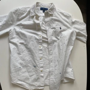 Vit kortärmad skjorta Ralph Lauren - Vit kortärmad skjorta från Ralph Lauren med blått broderat logotyp på bröstet. Skjortan har knappar framtill och är dekorerad med diskreta blå färgstänk för en cool, avslappnad look. Tillverkad i mjuk bomull och passar perfekt till jeans eller shorts.