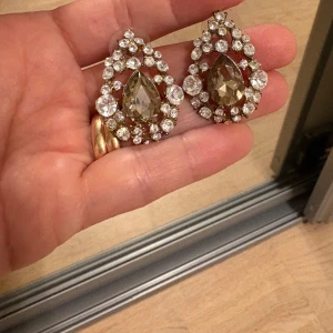  örhängen - Säljer ett par stora, droppformade örhängen med glittriga stenar i silverfärg och en stor brun sten i mitten. Perfekta för att ge din look extra bling! Stenarna är infattade i en snygg ram av mindre kristaller som verkligen fångar ljuset. Använd 1 gång
