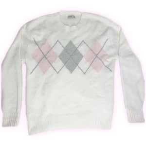 Vit argyle stickad tröja Hollister - Vit stickad tröja från Hollister med klassiskt argylemönster i grått och ljusrosa över bröstet. Rund halsringning och ribbade muddar ger en clean och mysig vibe. Perfekt för dig som gillar preppy och stilren look.