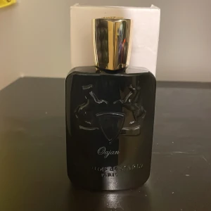 Parfums de Marly Oajan EdP 125ml - Lyxig parfymflaska i svart glas med guldfärgat lock från Parfums de Marly. Oajan har noter av honung, kanel, osmanthus, amber, labdanum, benzoin, vanilj, tonkaböna, patchouli och davana. Flaskan rymmer 125 ml Eau de Parfum och har elegant reliefdekor med två hästar. (Ungefär cirka 110 ml kvar, parfymen är en tester)