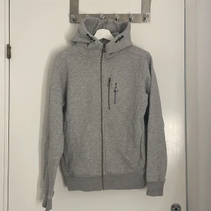 Grå hoodie från Sail Racing - Snygg grå hoodie från Sail Racing med dragkedja hela vägen, huva med snörning och en bröstficka med logga. Mjuk insida och ribbade muddar. Perfekt för dig som gillar en clean och sportig stil.