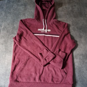 Vinröd hoodie från Jack & Jones - Snygg vinröd hoodie från Jack & Jones med vit snörning i huvan och tryckt logga på bröstet. Tröjan har en klassisk känguruficka och ribbade muddar. Perfekt för dig som gillar en avslappnad och stilren look. Storlek M men passar bra till S 