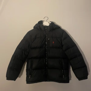 Polo Ralph Lauren jacka - Svart dunjacka från Polo Ralph Lauren med huva och dragkedja framtill. Jackan har två fickor med dragkedja och den klassiska röda Polo-loggan på bröstet. Puffig modell som håller dig varm under kalla dagar. Perfekt för dig som gillar stilren och sportig look. Den har små hål på ärmarna och 1 litet i nacken. Luvan gick av från sin dragkedja men är sydd tillbaka så det syns inte när man har på sig den. Pris kan diskuteras.
