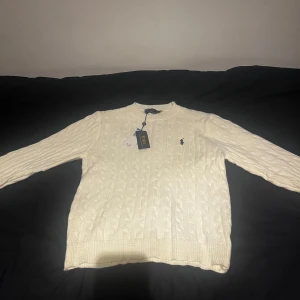 Vit kabelstickad tröja från Polo Ralph Lauren - Kabelstickad vit tröja från Polo Ralph Lauren med klassisk rund hals och broderad logga på bröstet. Tröjan har lång ärm och ribbade muddar för en snygg passform. Perfekt för dig som gillar stilren och tidlös design.