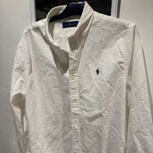 Vit skjorta från Ralph Lauren - Klassisk vit skjorta från Ralph Lauren med broderad blå logga på bröstet. Skjortan har lång ärm, knappar framtill och en stilren krage. Perfekt för dig som gillar en clean och tidlös look.för fler frågor eller bilder är de bara att skriva! 