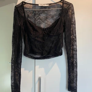 Svart spetsblus från NA-KD - Säljer en supersnygg helt oanvänd svart topp från NA-KD i transparent spets med blommigt mönster som tyvärr är för liten för mig! . Den har knappar framtill och croppad passform. Perfekt för dig som vill ha en trendig och snygg look.