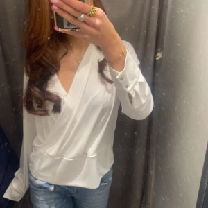 Vit omlottblus med v-ringning - Säljer en stilren vit omlottblus med v-ringning och långa ärmar. Blusen har knappar vid ärmsluten och ett snyggt omlottfall framtill som ger en elegant look. Perfekt att matcha med jeans eller kjol.