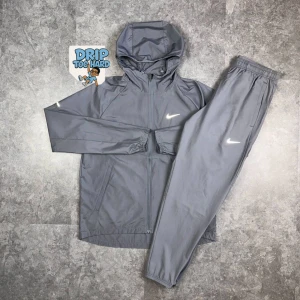 Grå träningsset från Nike - Snyggt grått träningsset från Nike med jacka och matchande byxor. Jackan har huva, dragkedja och två fickor, samt Nike-logga på bröstet. Byxorna har resår i midjan och logga på benet. Perfekt för dig som gillar sportig stil.