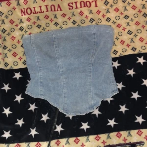 Ljusblå jeanskorsett med snörning - Säljer en snygg ljusblå korsett i jeanstyg med cool snörning framtill. Toppen har en markerad midja och en något längre, böjd nederkant. Perfekt att styla med kjol eller byxor för en trendig look.